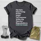 Dark gray t-shirt with gymnast uneven bars skills text on a light wooden surface with socks and a drawstring bag.