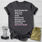Dark gray t-shirt with balance beam gymnast moves text on a light wooden surface with socks and a drawstring bag.