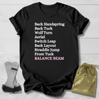 Black t-shirt with gymnast balance beam  moves text on a wooden surface with socks and a drawstring bag.