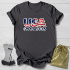 Gray t-shirt with 'USA Gymnastics' design on a light wooden surface, with socks and a drawstring bag in the foreground.