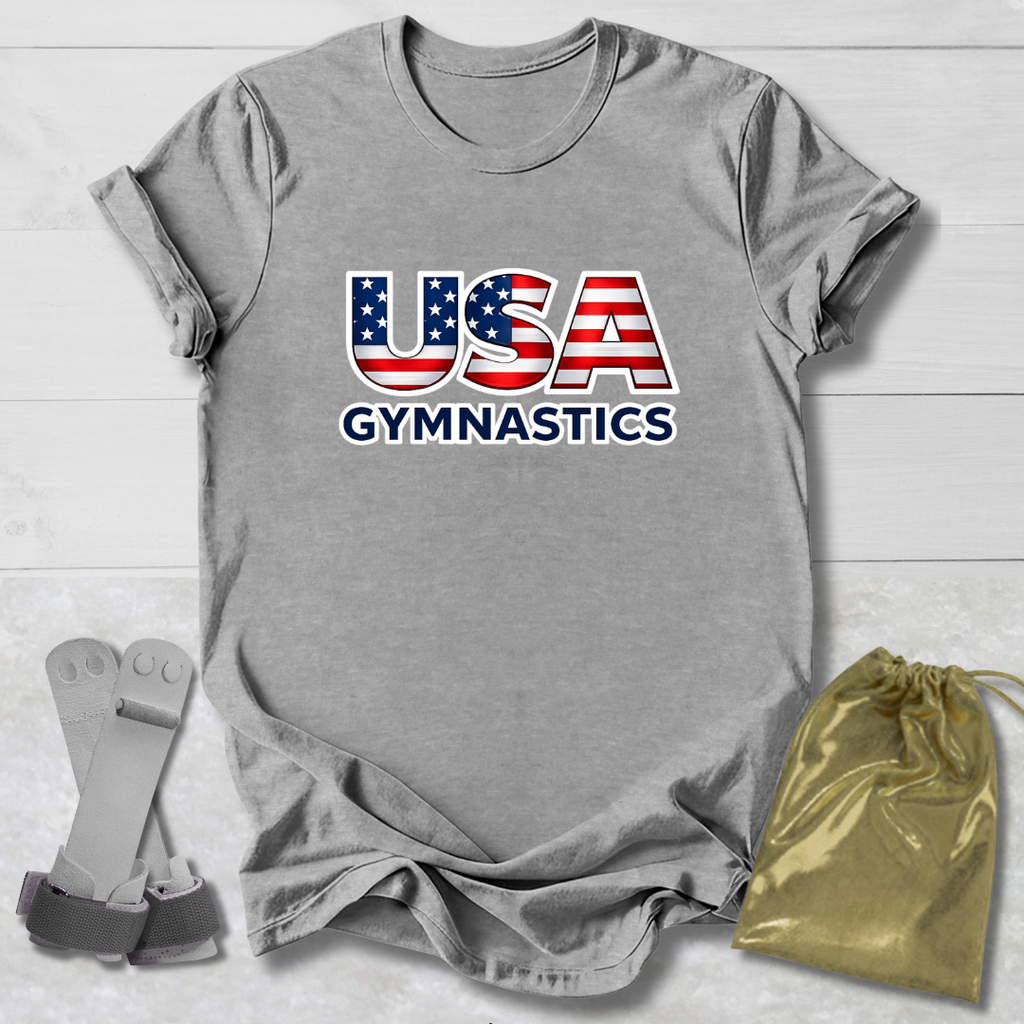 Gray t-shirt with 'USA Gymnastics' design on a wooden surface, accompanied by gray socks and a gold drawstring bag.