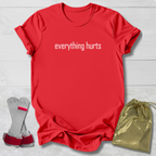 Red t-shirt with 'everything hurts' text, gray socks, and gold drawstring bag on a light wooden surface.