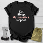 Black t-shirt with 'Eat. Sleep. Gymnastics. Repeat.' text on a light wooden surface with socks and a drawstring bag.