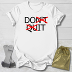 White t-shirt with 'DON'T QUIT' text on a light wooden surface with socks and a drawstring bag.
