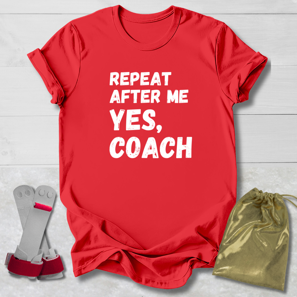 Red t-shirt with 'Repeat After Me Yes, Coach' text on a light wooden surface with socks and a drawstring bag.