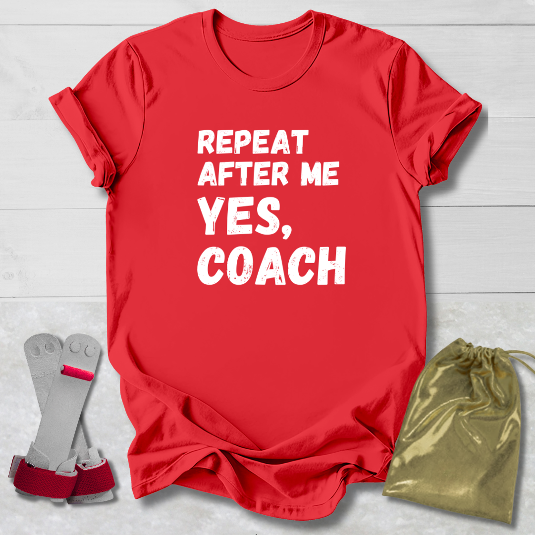 Red t-shirt with 'Repeat After Me Yes, Coach' text on a light wooden surface with socks and a drawstring bag.
