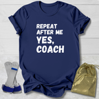 Navy blue t-shirt with 'Repeat after me yes, coach' text on a light wooden surface with socks and a drawstring bag.