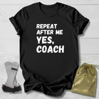 Black t-shirt with 'Repeat after me, yes, coach' text on a wooden surface with socks and a drawstring bag.