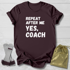 Maroon t-shirt with 'Repeat after me yes, coach' text on a light wooden surface with socks and a drawstring bag.