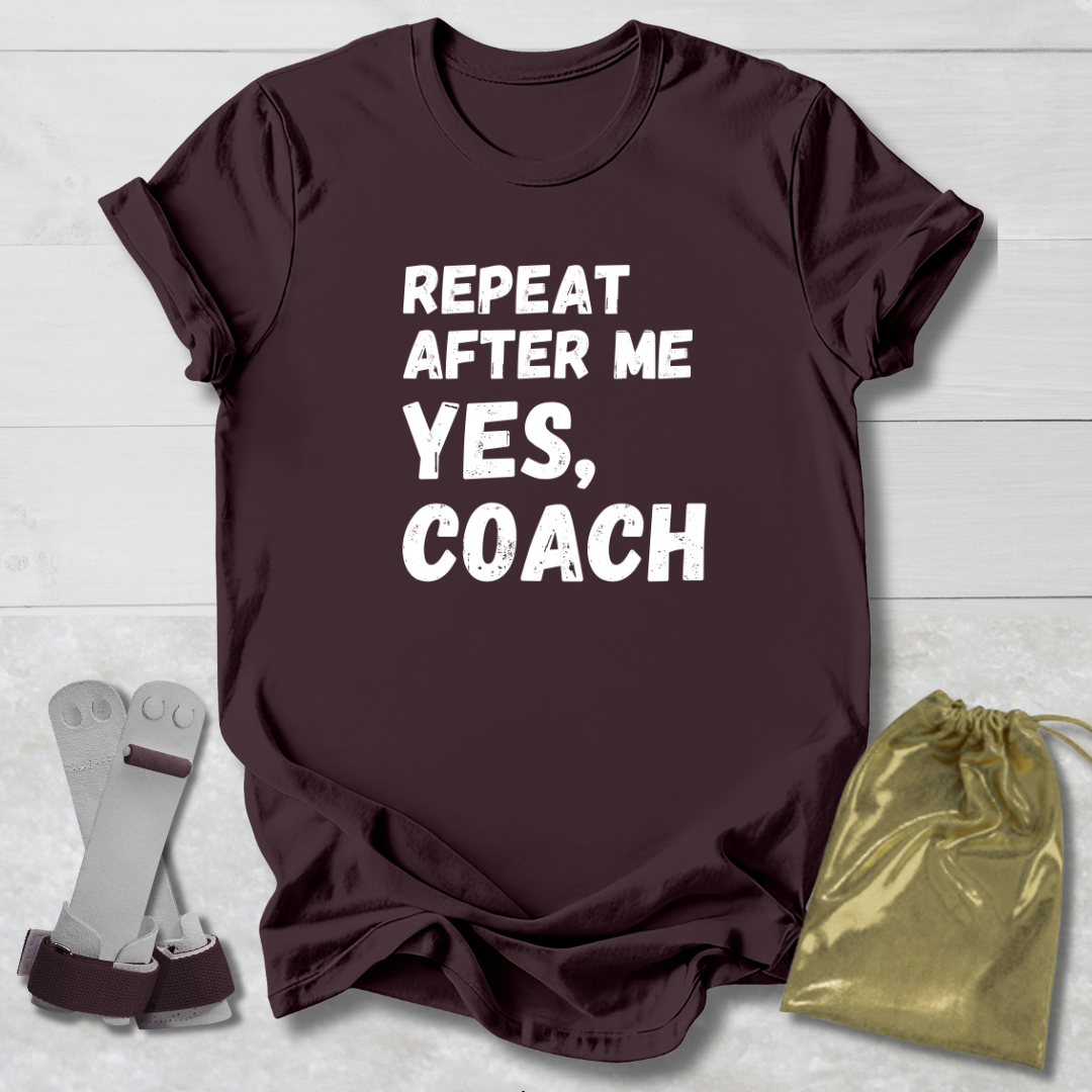 Maroon t-shirt with 'Repeat after me yes, coach' text on a light wooden surface with socks and a drawstring bag.