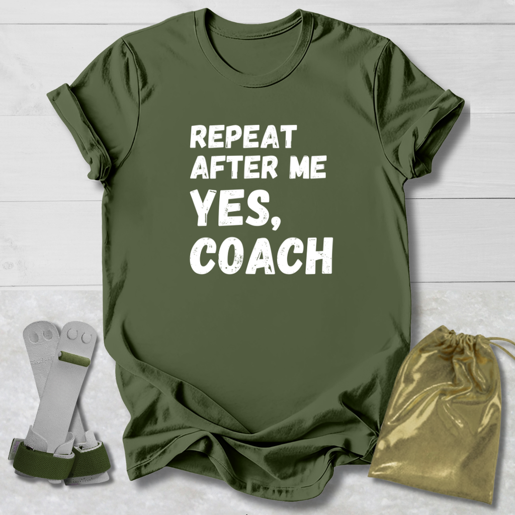 Green t-shirt with 'Repeat after me yes, coach' text on a wooden surface with socks and a drawstring bag.