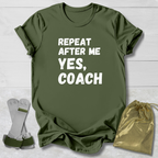 Green t-shirt with 'Repeat after me yes, coach' text on a wooden surface with socks and a drawstring bag.
