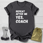 Gray t-shirt with 'Repeat after me, yes, coach' text on a light wooden surface with socks and a drawstring bag.