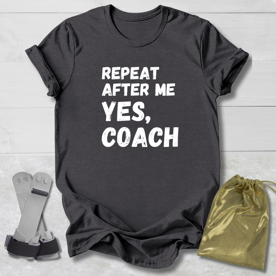 Gray t-shirt with 'Repeat after me, yes, coach' text on a light wooden surface with socks and a drawstring bag.