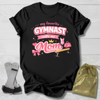 Black t-shirt with pink gymnastics-themed design on a light wooden surface.
