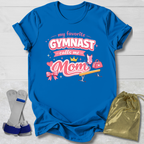 Blue t-shirt with 'My favorite gymnast calls me Mom' text, socks, and drawstring bag on a light wooden surface.