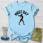 Light blue t-shirt with 'MEET DAY' text and silhouette, socks, and drawstring bag on a wooden surface.