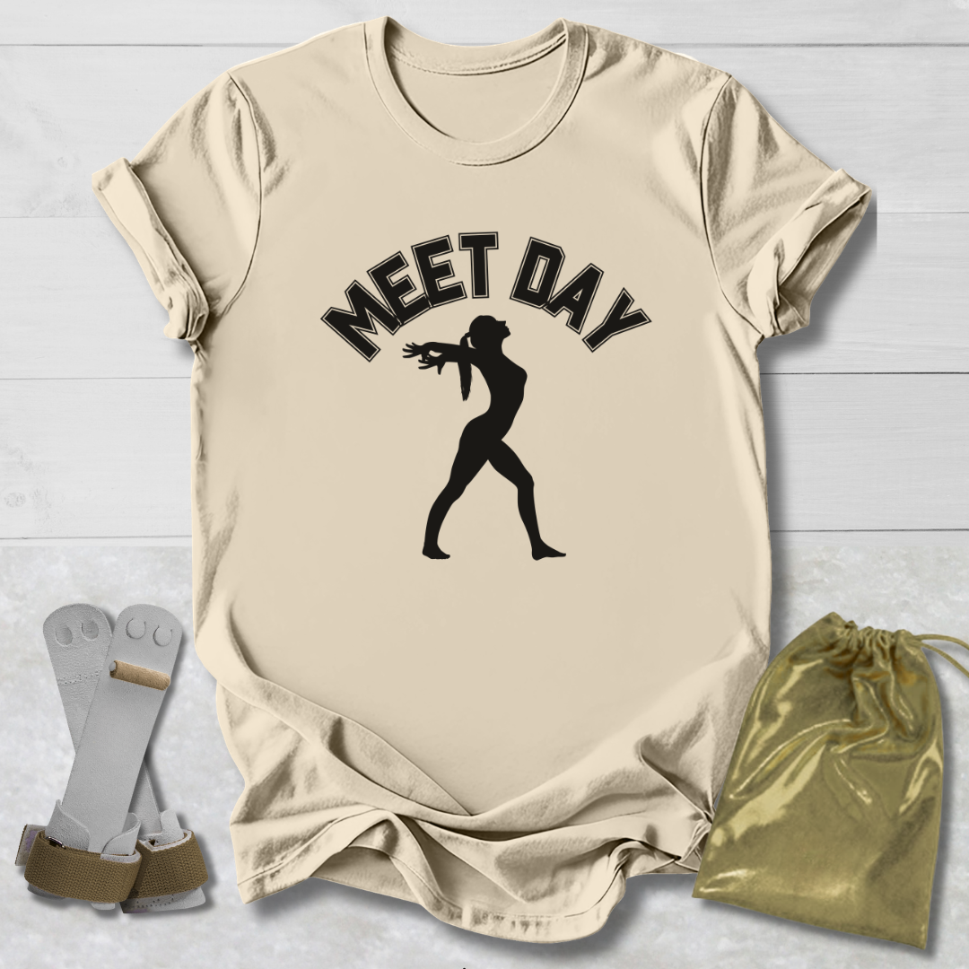 Beige t-shirt with black gymnast graphic and Meet Day text, socks, and drawstring bag on a light wooden surface