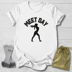 White t-shirt with black gymnast graphic and Meet Day text, socks, and drawstring bag on a light wooden surface.