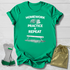 Green t-shirt with 'Homework Practice Repeat' text and graphics, socks, and drawstring bag on a light wooden surface.
