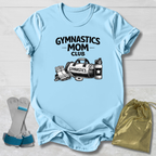 Light blue t-shirt with 'Gymnastics Mom Club' text and graphics on a wooden surface, with socks and a drawstring bag in the foreground.