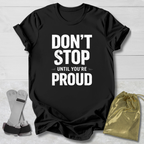 Black t-shirt with 'Don't Stop Until You're Proud' text on a light wooden surface with socks and a drawstring bag.