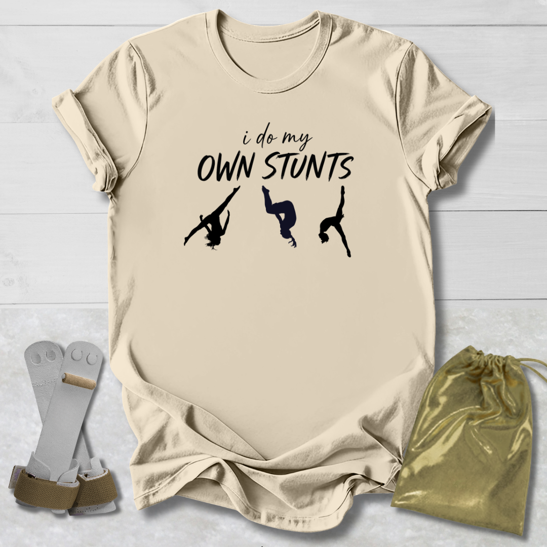 Beige t-shirt with 'i do my own stunts' text and silhouettes, socks, and drawstring bag on a light wooden surface.
