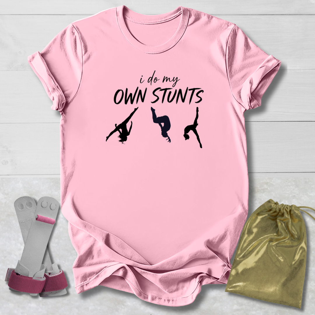 Pink t-shirt with 'i do my own stunts' text and silhouettes, socks, and drawstring bag on a light wooden surface.
