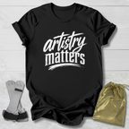 Black t-shirt with 'artistry matters' text on a light wooden surface with socks and a drawstring bag.