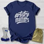 Navy blue t-shirt with 'artistry matters' text, socks, and a drawstring bag on a light wooden surface.
