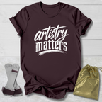 Maroon t-shirt with 'artistry matters' text, socks, and a drawstring bag on a light wooden surface.