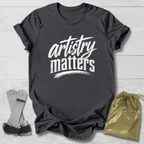 Dark gray t-shirt with 'artistry matters' text, socks, and a drawstring bag on a light wooden surface.