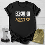 Black t-shirt with 'Execution Matters' text on a wooden surface with socks and a drawstring bag.