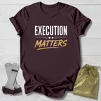 Maroon t-shirt with 'EXECUTION MATTERS' text on a light wooden surface with socks and a drawstring bag.