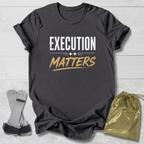 Dark gray t-shirt with 'Execution Matters' text on a light wooden surface with socks and a drawstring bag.