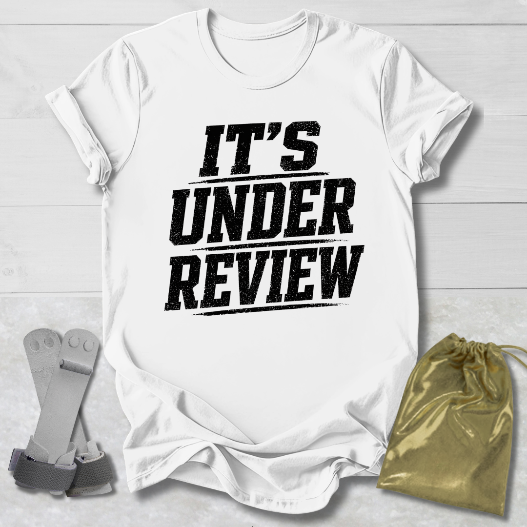 White t-shirt with 'IT'S UNDER REVIEW' text on a light wooden surface with socks and a drawstring bag.
