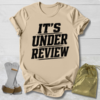 Beige t-shirt with 'IT'S UNDER REVIEW' text, socks, and drawstring bag on a light wooden surface