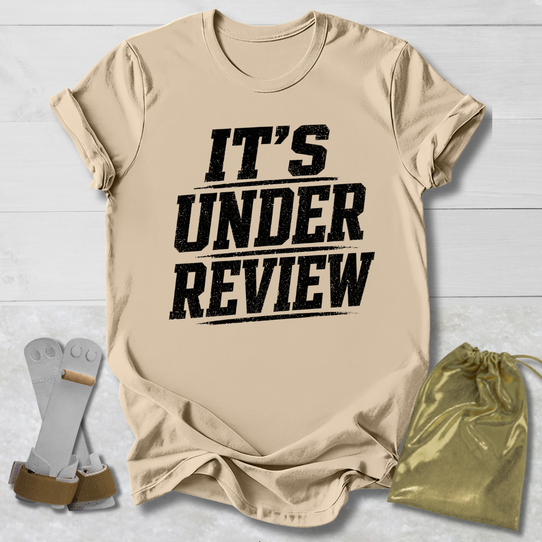 Beige t-shirt with 'IT'S UNDER REVIEW' text, socks, and drawstring bag on a light wooden surface