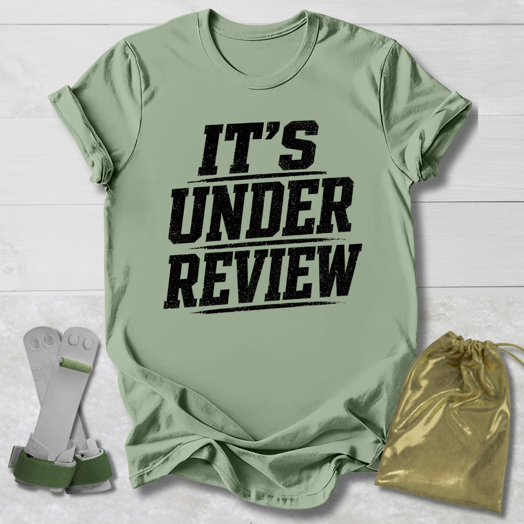Green t-shirt with 'IT'S UNDER REVIEW' text, socks, and drawstring bag on a light wooden surface.