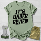 Green t-shirt with 'IT'S UNDER REVIEW' text, socks, and drawstring bag on a light wooden surface.