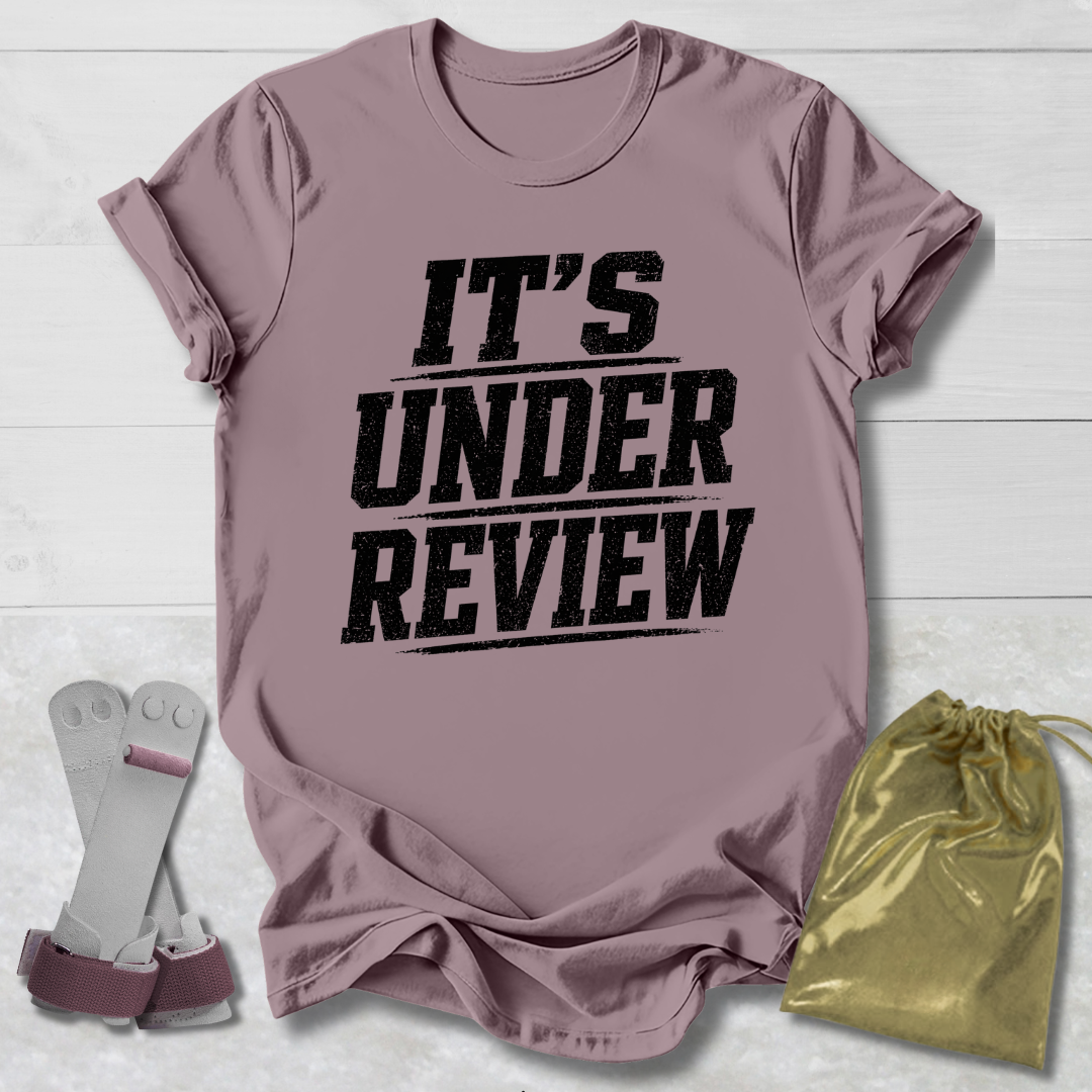 Mauve t-shirt with 'IT'S UNDER REVIEW' text, socks, and drawstring bag on a light wooden surface