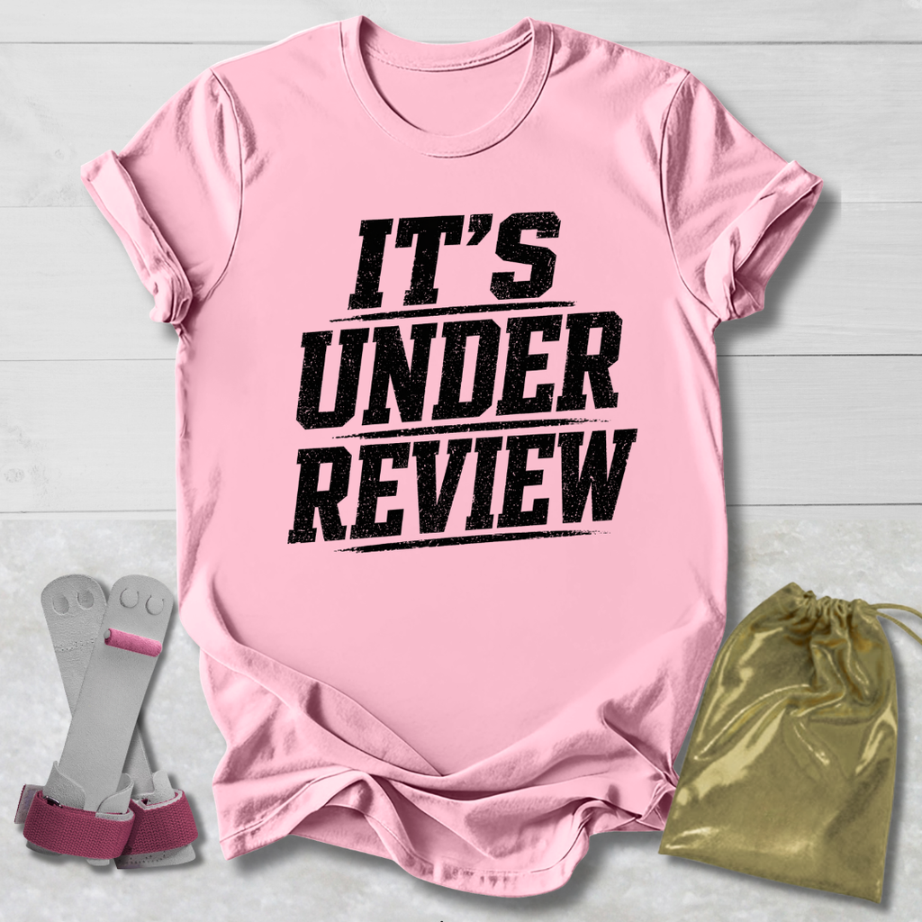 Pink t-shirt with 'IT'S UNDER REVIEW' text, socks, and drawstring bag on a light wooden surface.