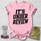 Pink t-shirt with 'IT'S UNDER REVIEW' text, socks, and drawstring bag on a light wooden surface.