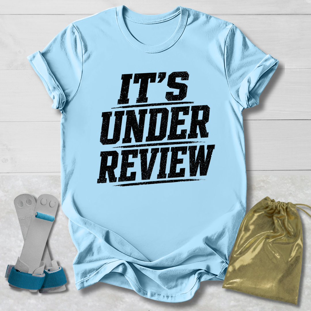 Light blue t-shirt with 'IT'S UNDER REVIEW' text, socks, and drawstring bag on a light wooden surface.