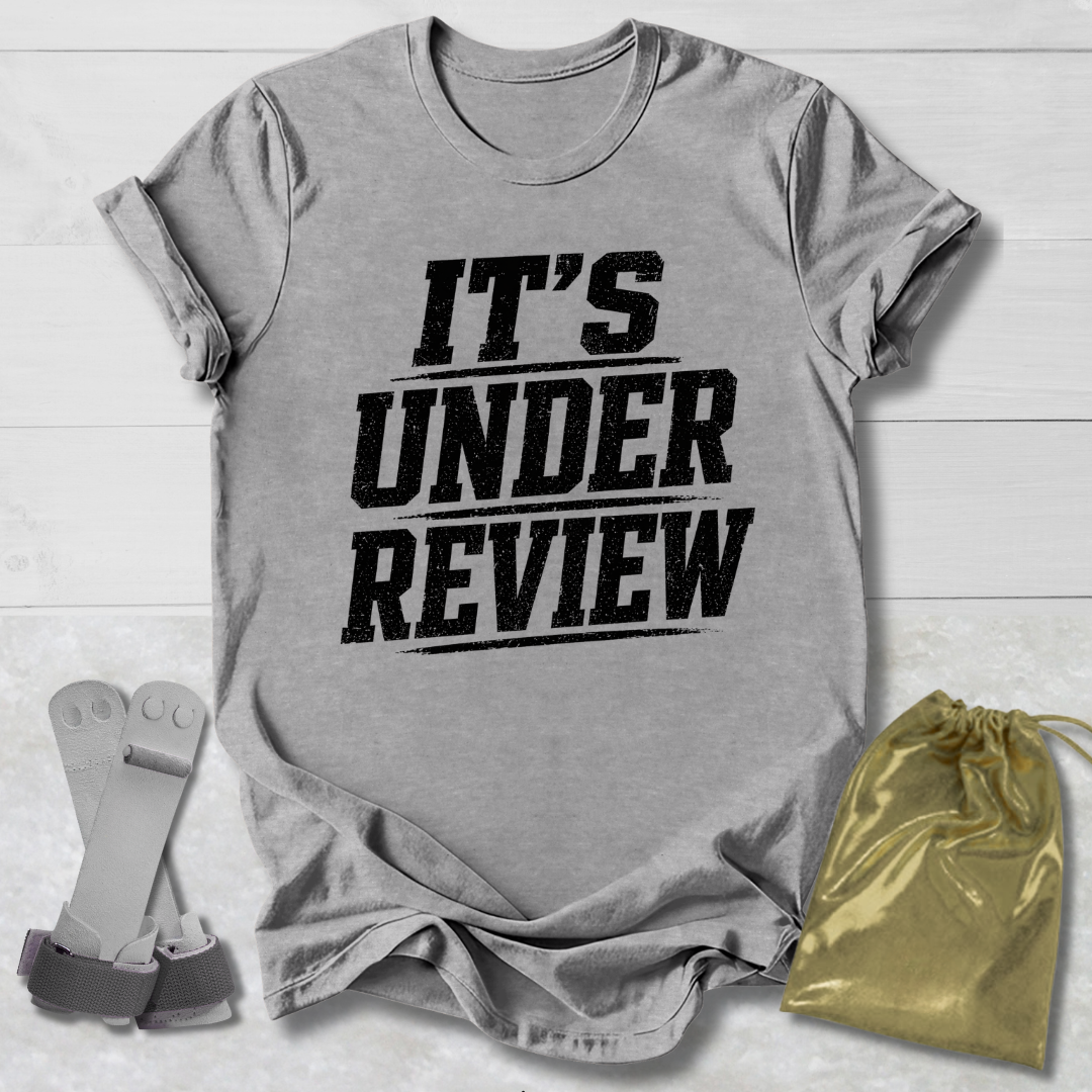 Gray t-shirt with 'IT'S UNDER REVIEW' text, socks, and drawstring bag on a light wooden surface