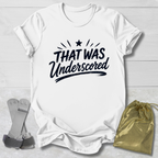 White t-shirt with 'THAT WAS Underscored' text on a light wooden surface with socks and a drawstring bag.