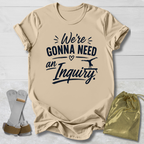 Beige t-shirt with text 'We're Gonna Need an Inquiry' on a light wooden surface with socks and a drawstring bag.