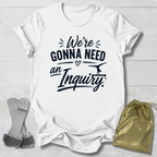 White t-shirt with black text 'We're Gonna Need an Inquiry' on a light wooden surface with socks and a drawstring bag.