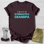 Maroon t-shirt with 'Proud Gymnastics Grandpa' text, socks, and drawstring bag on a light wooden surface.