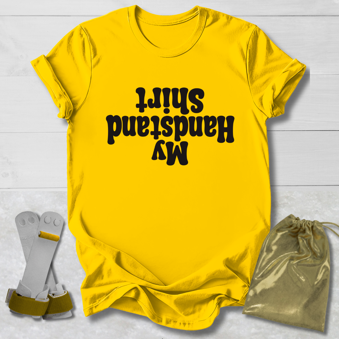 Yellow t-shirt with ' My Handstand Shirt' text on a light wooden surface with socks and a drawstring bag.